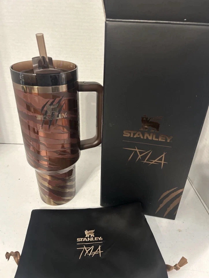 A Stanley Tyla insulated tumbler with a handle and straw, shown next to its black branded box and a black drawstring pouch on a light surface.