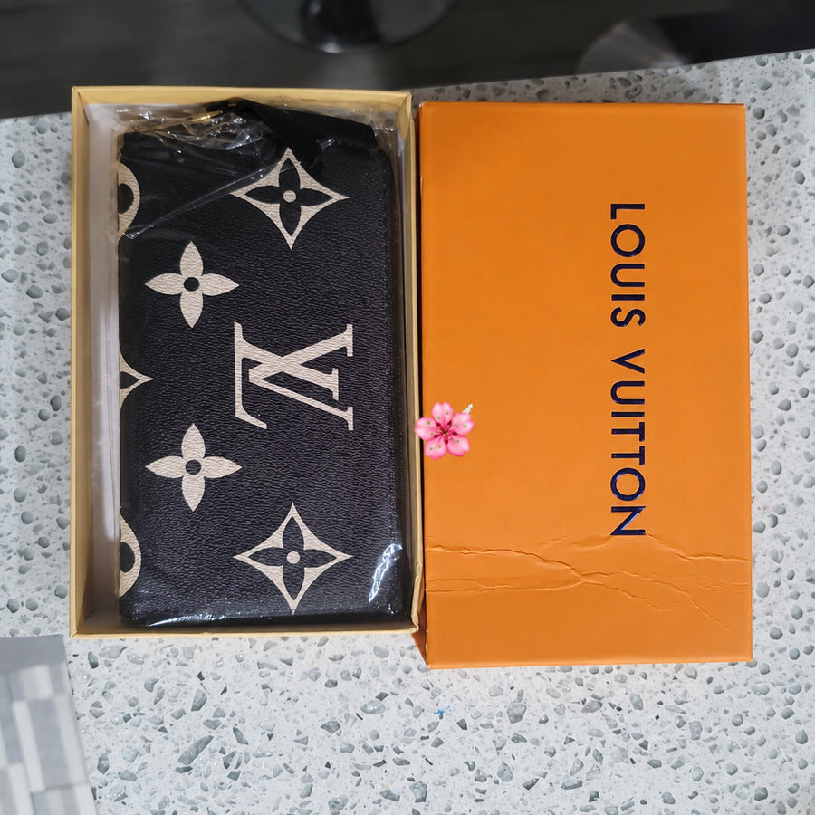A Louis Vuitton monogram wallet or card holder in dark brown or black with beige monogram motifs, shown inside an open Louis Vuitton box next to an orange Louis Vuitton box on a speckled countertop.