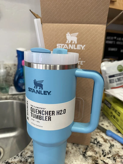 A light blue Stanley Quencher H2.0 Tumbler with a handle, lid, and straw, shown in front of its brown cardboard packaging box on a kitchen countertop.