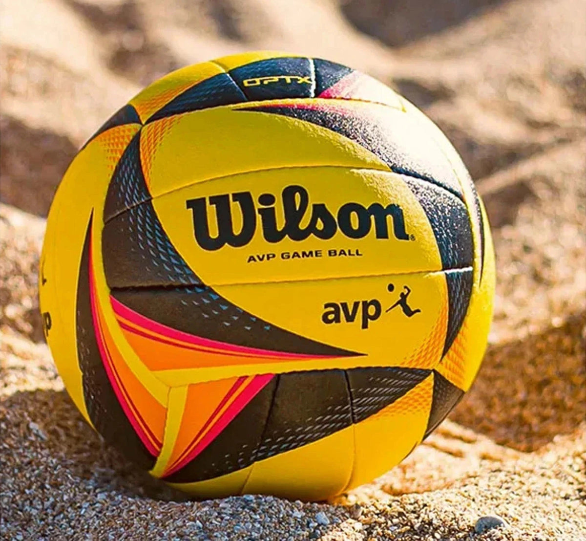 A Wilson AVP game ball volleyball resting on sand, showing its yellow, black, and orange panels with the Wilson logo.