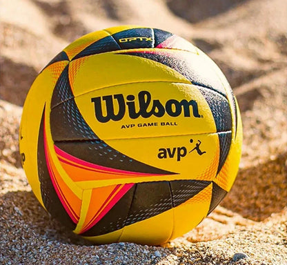 A Wilson AVP game ball volleyball resting on sand, showing its yellow, black, and orange panels with the Wilson logo.