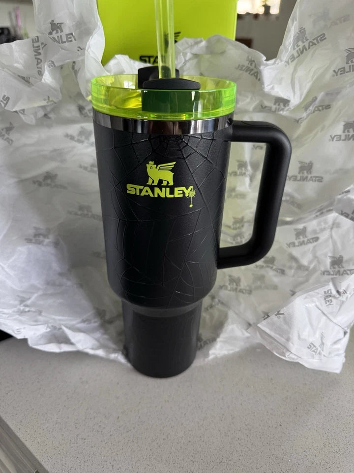 A black Stanley travel tumbler with a handle and lid, featuring a bright yellow-green logo and cracked-glass style texture, set on a countertop with branded tissue paper around it.