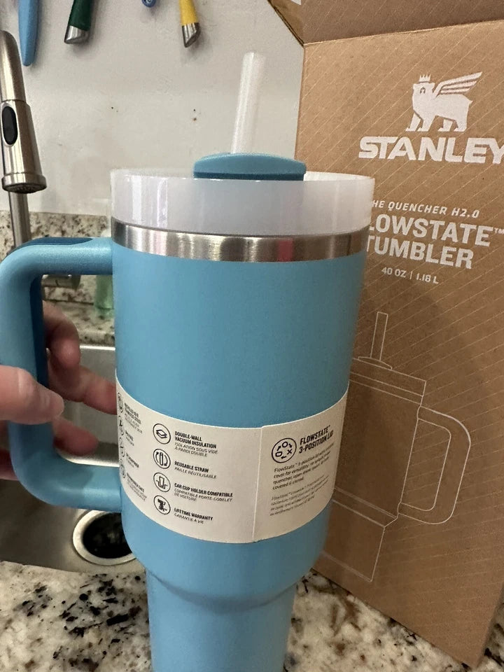 A light blue Stanley tumbler with a handle, clear lid, and straw, held by a hand in front of its brown branded packaging box on a kitchen countertop.
