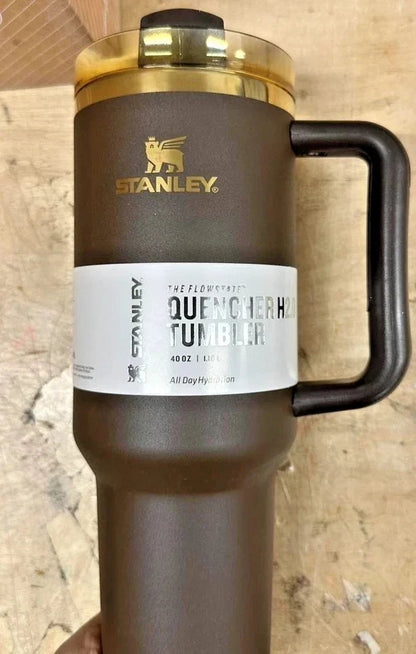 Stanley Quencher H2 Tumbler in matte dark brown with a black handle, gold logo, and a white paper label on a textured beige background.