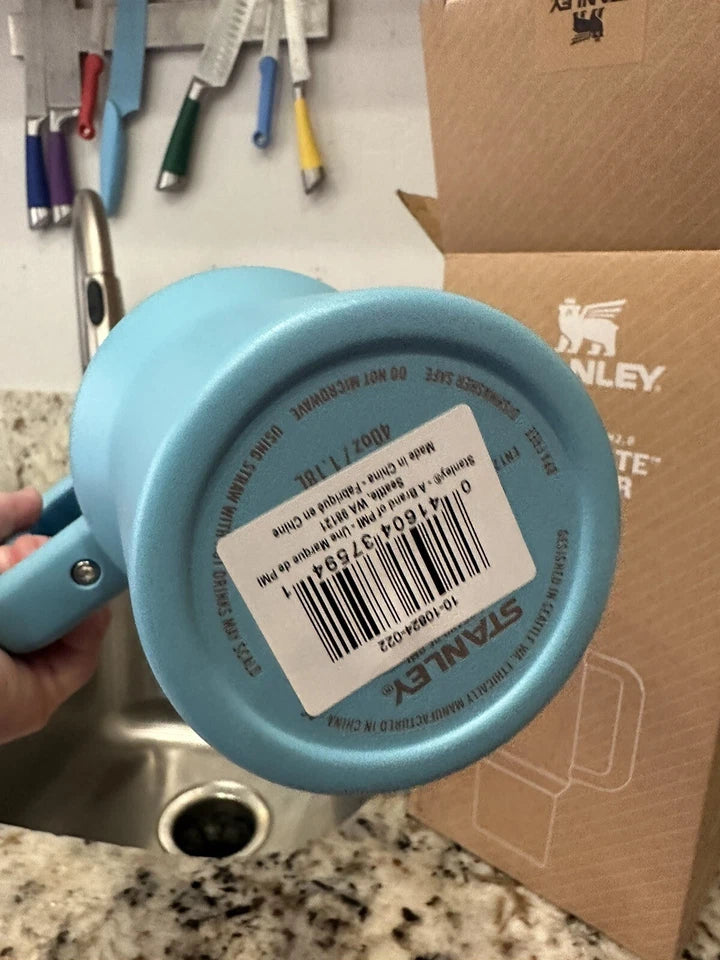A light blue Stanley mug is held by a hand, showing the bottom of the mug with a barcode sticker and Stanley branding. A cardboard Stanley box is visible in the background on a kitchen counter.